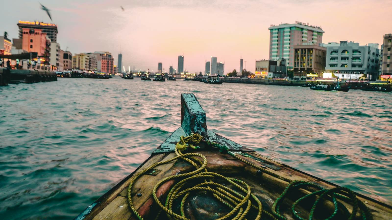 The bow of a wooden abra crossing Dubai Creek at dusk, with the waterfront and the city's towers ahead.