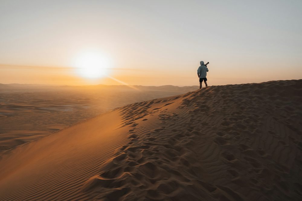 A person standing on the crest of a sand dune as the sun sets over the desert.